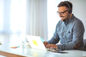 SE Telecom Canadian professional smiling while using a telephone for VoIP softphone solution on laptop with headset at his desk